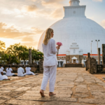 Une voyageuse blonde en vêtements de lin blanc marche pieds nus vers un grand stupa blanc dagoba pendant lheure dorée tenant une offrande de fleur de lotus pendant que des dévots locaux prient