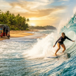 Une photographie candide au téléobjectif capturant une surfeuse surfant sur une puissante vague tubulaire turquoise et vitreuse Des embruns brillants sont figés dans lair autour delle Loin en arrière plan considérablement floue et plus petite se trouve la plage sri lankaise avec le groupe de personnes et le tuk tuk rouge et jaune avec des planches de surf sur le toit La scène est baignée par la riche et chaude lueur dorée dun coucher de soleil