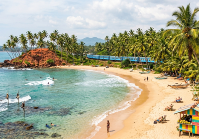 Vue panoramique d'une plage tropicale au Sri Lanka avec du sable doré, des cocotiers sur une colline rouge, des pêcheurs sur échasses et l'océan Indien turquoises.